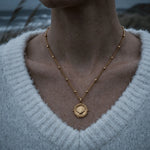 The Mermaid's Coin - a 2cm round gold disc pendant featuring a bold seashell motif and the Latin inscription 'Fluctuat nec mergitur', handcrafted by Dainty London.